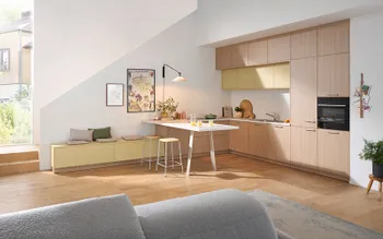 Helle Wohnküche mit Holzfronten, gelben Oberschränken und weißem Frühstückstresen unter einer schrägen Wand mit Sitzbank und Dekoration.